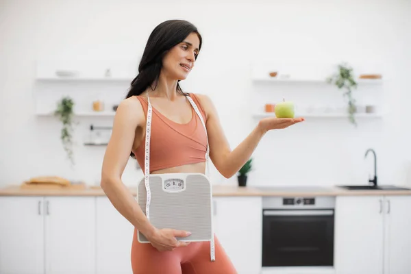 Slender caucasian woman with measuring tape and scales holding apple on palm while standing in kitchen. Effective health coach in sportswear assisting in weight management via healthy nutrition.