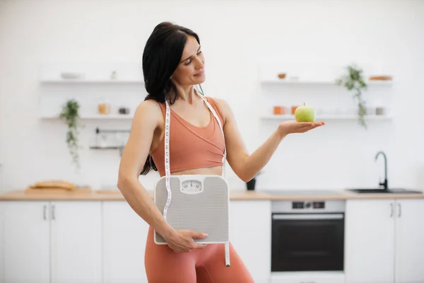 Slender caucasian woman with measuring tape and scales holding apple on palm while standing in kitchen. Effective health coach in sportswear assisting in weight management via healthy nutrition.