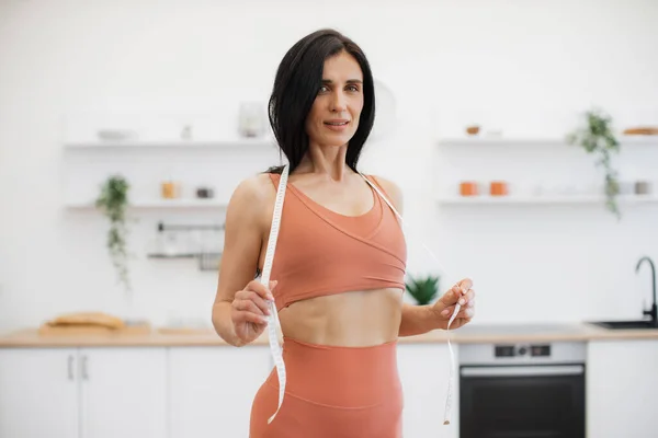 Portrait of skinny woman in terracotta gym clothes wearing measuring tape around neck in modern kitchen. Sport-loving person developing healthy habits through diet and daily exercise at home.