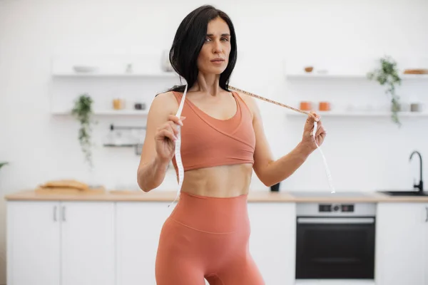 Portrait of skinny woman in terracotta gym clothes wearing measuring tape around neck in modern kitchen. Sport-loving person developing healthy habits through diet and daily exercise at home.