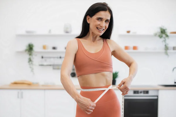 穿着瑜伽运动服在家锻炼后 迷人的女性在健康的腰围上用胶带测量 运动员通过饮食习惯和健身来引导人们朝着改善福祉的方向前进 — 图库照片