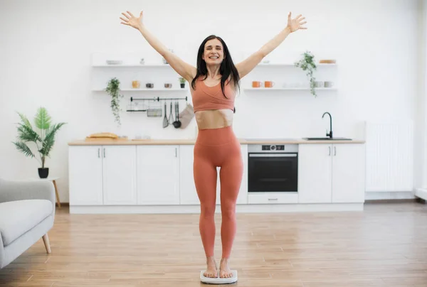 Full length view of woman in stylish activewear raising arms in victory gesture while standing on bath scales at home. Vigorous caucasian lady celebrating achievements in healthy lifestyle changes.