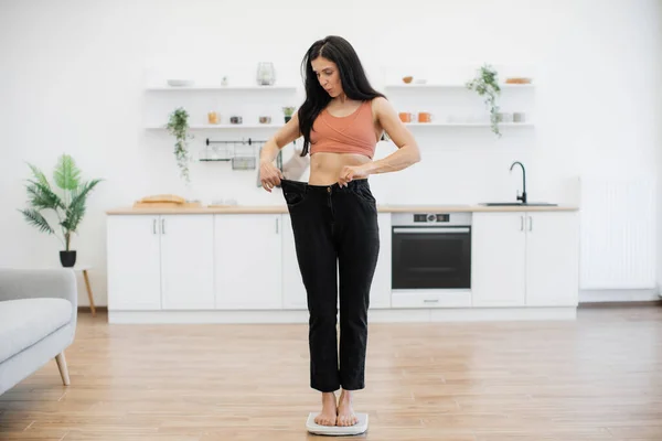 現代のキッチンの真ん中にスケールに立っている間 スリムな姿のゆったりとしたズボンを見つめている驚くべきブルネットの女性 栄養療法で達成された成功の楽しいショックを感じる幸せな女性 — ストック写真