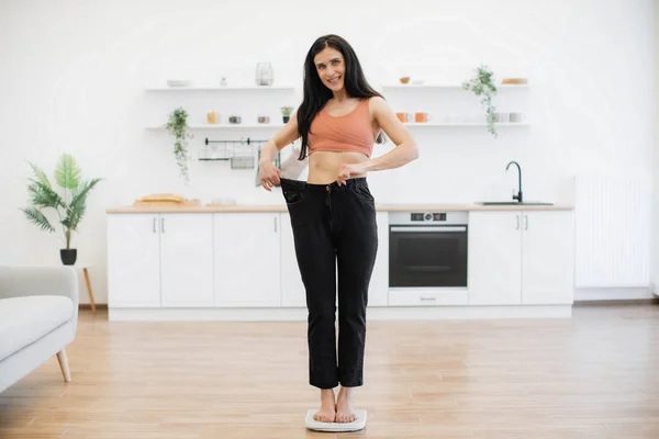 Amazed brunette female staring at loose pants on slim figure while standing on scales in middle of modern kitchen. Happy woman feeling pleasant shock of attained success in nutritional therapy.