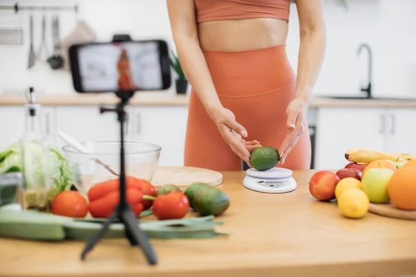 スポーティな女性が三脚の携帯電話の後ろのテーブルに立っている間 栄養スケールにアボカドを置くという風景 健康ブログのために活発に食べることについてのビデオコンテンツを撮影スリムフィットネスコーチ — ストック写真