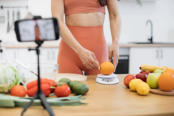 スマートデジタルスケールでオレンジを測定するスレンダー女性のクローズアップビューは プロセスを撮影する際のガジェットです 良い形の女性は 低カロリー製品で食事療法を改善するためのアドバイスを与える — ストック写真