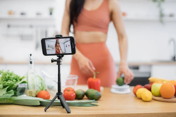 デジタル画面でアボカドを評価するスレンダー女性のイメージを持つ三脚が保有する携帯電話のクローズアップビュー キッチンインテリアで新鮮なベジタリアンサラダの食材を準備するフードブロガー — ストック写真