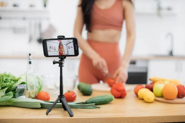 ブルネットの女性スライストマトのカッティングボードのクロップビューとモバイルでのプロセスの記録を得る アクティブウェアのアスレチック成人 栄養豊富な野菜を介して食事の変更についてブログ — ストック写真