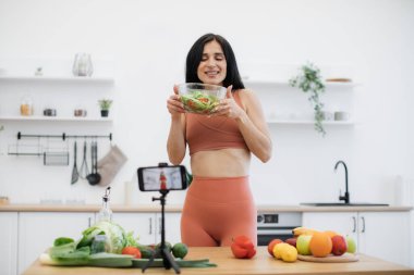 Durakta şık bir akıllı telefon spor elbiseli mutlu bir kadını besin tabağı ile gösteriyor. Çekici esmer kadın abonelerle yemek pişirmenin zevkini ve neşesini paylaşıyor..