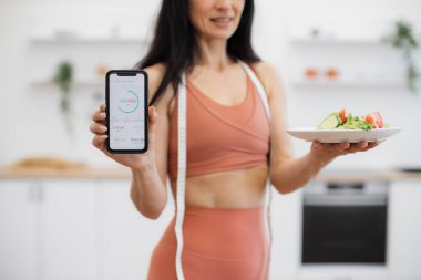 Akıllı telefon ve taze salatayla poz veren ve ekranda tüketilen kalori miktarını gösteren spor giysili kadın ürünleri. Enerjik bir kadın günlük beslenme planı için spor takip cihazı kullanıyor..