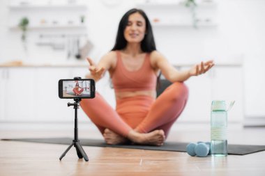 Profesyonel bayan antrenör evdeki fiziksel aktivite sırasında tripod ve akıllı telefon kullanarak video kaydediyor. Nilüfer çiçeğinin üzerinde otururken meditasyon yapan ve kameraya gülümseyen kadının bulanık arkaplanı..