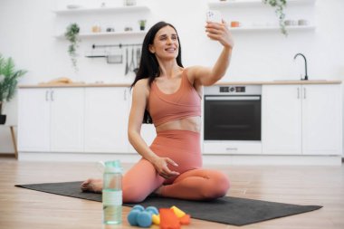 Sıska esmer kadın, beyaz ferah mutfakta lastik minder üzerinde çalışırken akıllı telefondan selfie çekiyor. Spor kıyafetleri ve spor malzemeleri eğitimi alan güzel bir kadın..
