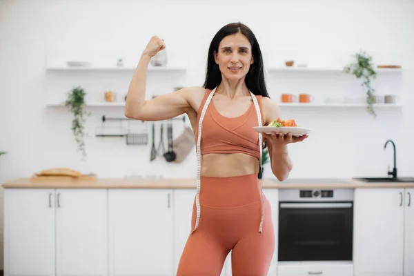 Beautiful sporty woman showing arm muscles while standing in bright trendy kitchen with vegetables plate. Portrait of strong female dressed in terracotta sportswear posing at camera with healthy food.