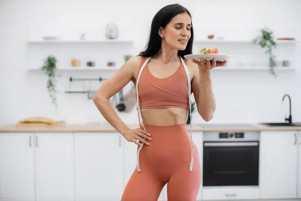 Pretty brunette woman enjoying smell of healthy organic salad while standing in kitchen after regular workout. Athletic female with prefect body shape consuming low calorie food to avoid weight gain.