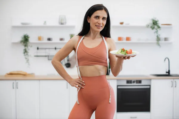 Portrait of fit and slender woman standing in middle of modern white kitchen and looking at camera. Attractive woman in terracotta tight sport outfit posing with fresh salad and gently smiling.