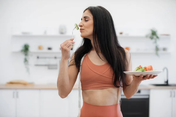 自宅で効率的なトレーニングの後 低カロリーの食べ物を食べながら新鮮なキュウリの匂いを楽しんでいるエネルギッシュフィットネス女性 有機食を摂取して健康を守るアクティブな女性 — ストック写真