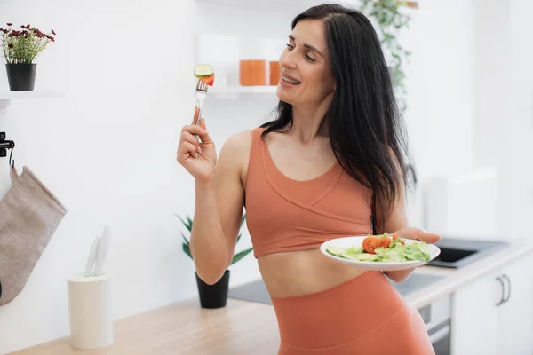 スリムな慎重なブルネット女性は タイトなレギンスを着て キッチンに立って脇を見ながら健康的なビーガンサラダを楽しんでいます フィットネスと健康的なライフスタイルのコンセプト — ストック写真