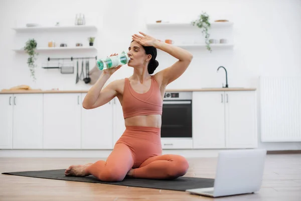 stock image Slim woman dressed in tight leggings and top feeling thirsty after intense workout and drinking fresh water. Sporty lady with hair gathered in bun exercising on yoga mat and enjoying music on laptop.