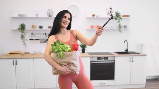 穿着运动服的迷人女人用手机把新鲜产品装进麻袋 然后粘在家里 通过体育活动和适当饮食保持健康体重的瘦小女性 — 图库视频影像