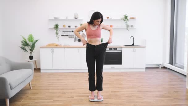 現代のキッチンの真ん中にスケールに立っている間 スリムな姿のゆったりとしたズボンを見つめている驚くべきブルネットの女性 栄養療法で達成された成功の楽しいショックを感じる幸せな女性 — ストック動画