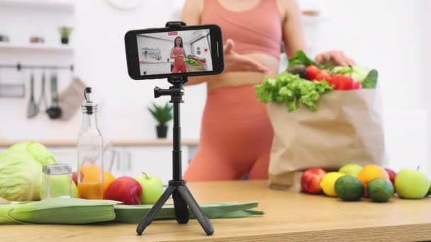 专注于工作相机应用的智能装置 由三脚架固定在厨房桌子上 背景为纤细女性 快乐的Vlogger与西红柿射击视频内容有关运动营养食品 — 图库视频影像