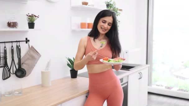 キッチンに立っているジム服を着て グリーンでプレートを見ている人 オーガニックなバランスの取れた食べ物と定期的なトレーニングで健康的なライフスタイルを促進する長髪ブルネット — ストック動画