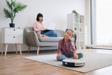 Genç bir bayan dizüstü bilgisayar kullanırken akıllı temizlik cihazının arkasında yerde oturan sevimli kızın tam boy görüntüsü. Mutlu anne ve kız akıllı aletlerle rahat bir yaşam sürüyorlar..