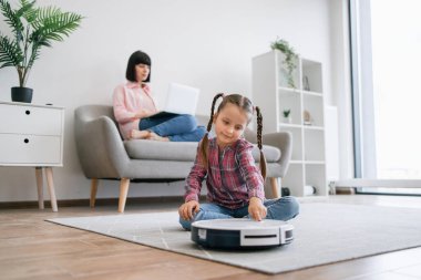 Genç bir bayan dizüstü bilgisayar kullanırken akıllı temizlik cihazının arkasında yerde oturan sevimli kızın tam boy görüntüsü. Mutlu anne ve kız akıllı aletlerle rahat bir yaşam sürüyorlar..