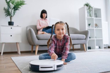Saçı sıkıca örülmüş bir kızın portresi yerde bağdaş kurarak otururken akıllı bir teknolojik aleti aktifleştiriyor. Akıllı ergen kız rutin temizliğe başlarken anne dizüstü bilgisayarında e-posta yazıyor..