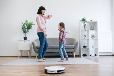 Robotik süpürge zemini düzgünce temizlerken küçük kıza dans hareketleri gösteren genç bayan. Ev sahipleri ev işleri için elektrikli aletleri kullanırken anne-kız partisi için zaman ayırıyorlar..