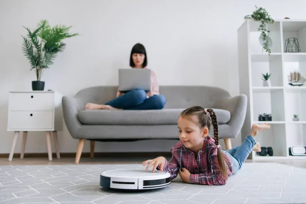 Little girl watching robot vacuum performing automatic cleaning while young woman with computer doing remote job. Smart technologies allowing mom and kid focusing on other tasks but household chores.