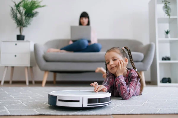 Focus on well-rounded robot vacuum cleaner being placed on floor of living room by cute child in casual clothes. Stay-at-home mom involving offspring to housekeeping duties while running business.