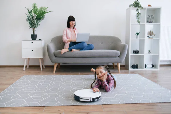 Focus on well-rounded robot vacuum cleaner being placed on floor of living room by cute child in casual clothes. Stay-at-home mom involving offspring to housekeeping duties while running business.