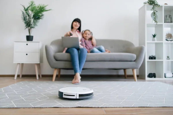 Focus on automatic cleaning robot controlled remotely by brunette woman with computer sitting near preteen girl. Happy mom and daughter starting daily vacuuming while having cinema day at home.