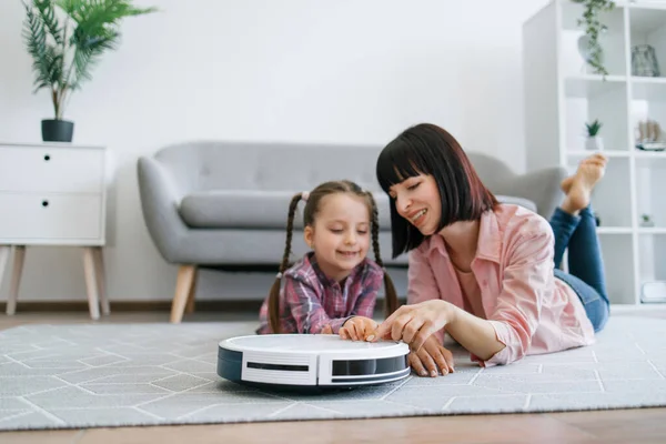 Close up view of joyful woman and tween girl activating robotic cleaner while resting on carpet in apartment. Delighted parent and child following rules of using smart electrical appliance at home.