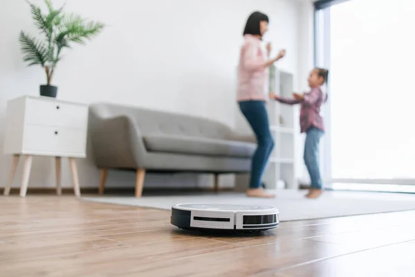 Intelligent cleaning machine collecting dust on wooden surface while two ladies in cozy outfits rocking out to tunes. Mother and daughter testing new playlist while robotic device getting area tidied.