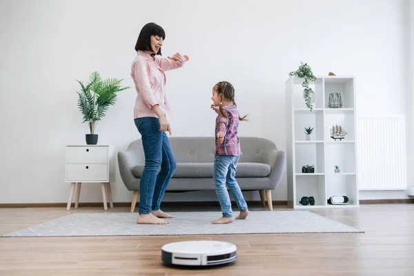 Young lady showing dance moves to little girl while robotic vacuum running floor smoothly. Homeowners using electrical appliance for household chores while freeing up time for mother-daughter party.
