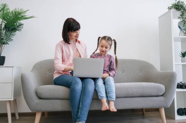 Oturma odasındaki koltukta modern dizüstü bilgisayarla poz veren kot pantolonlu alımlı genç bayanların portresi. Memnuniyetle aile üyeleri evdeki akıllı aygıtla eski dijital fotoğraf albümlerini araştırıyor..