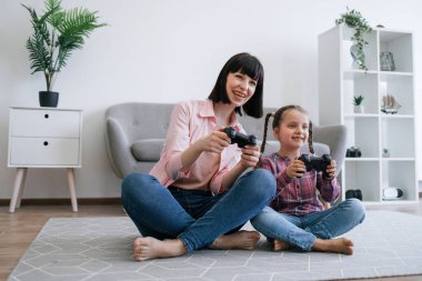 Bacak bacak üstüne atmış bir şekilde odada dinlenirken elinde siyah joypad 'ler tutan keyifli kadın ebeveyn ve kız. Yarışma ve bilgisayar eğitimi için video oyunu oynayan aşırı neşeli aile üyeleri.