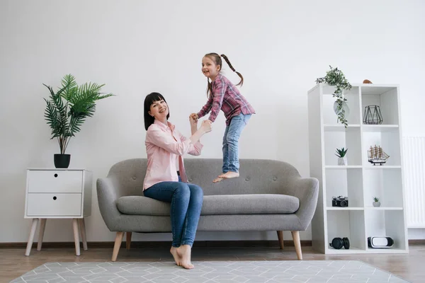 Full length view of smiling lady in jeans supporting cute girl in testing great bounce of cushiony couch in family lounge. Delighted mom-daughter duo experiencing feeling of freedom and well-being.