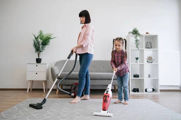 Brunette woman and tween girl utilizing different models of vacuum cleaners while removing dirt in living room. Diligent young homemakers sharing responsibilities of keeping apartment tidy.