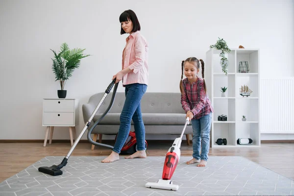 Brunette woman and tween girl utilizing different models of vacuum cleaners while removing dirt in living room. Diligent young homemakers sharing responsibilities of keeping apartment tidy.