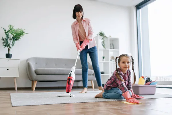 Preteen kid in latex gloves cleaning floor from dirt while mother removing dust from carpet with canister vacuum. Tireless ladies of house reducing risk of allergies by taking care of home chores.