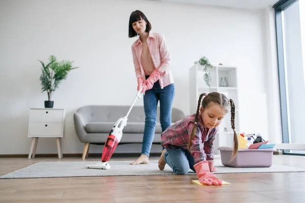 Helpful tween girl in rubber gloves polishing laminate floor while brunette woman vacuuming rug with electric appliance. Focused stay-at-home mom involving daughter in household chores at weekend.