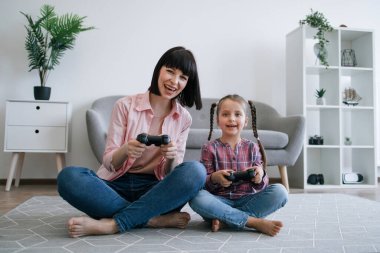Modern bir dairede halıda gülerken, elinde oyun konsolu tutan mutlu anne ve ergen kızın yakın görüntüsü. Oyuncu yetişkin ve çocuk boş zamanlarında sanal dünyanın zorluklarını kucaklıyor.