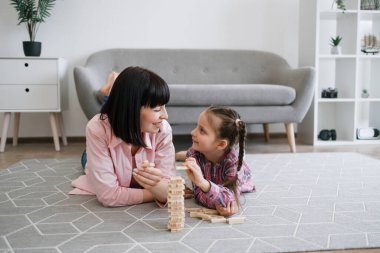 Neşeli beyaz kadın yetişkin ve çocuk ellerinde oyuncak bloklarla oturma odasının halısında dinleniyor. Sevecen anne ve kız, ahşap tuğlaları dizerken el-göz koordinasyonunu geliştiriyorlar..