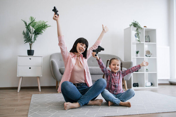 Emotional family ladies in denim clothes raising hands with joysticks in air while resting on carpet in lounge. Dedicated gamers expressing delight with victory gesture after successful final.