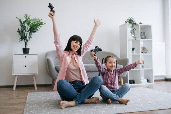 Emotional family ladies in denim clothes raising hands with joysticks in air while resting on carpet in lounge. Dedicated gamers expressing delight with victory gesture after successful final.