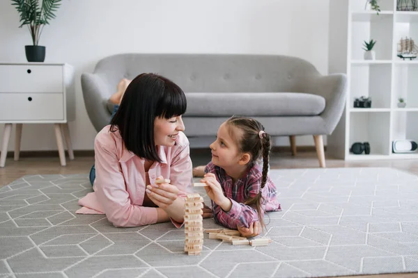 Neşeli beyaz kadın yetişkin ve çocuk ellerinde oyuncak bloklarla oturma odasının halısında dinleniyor. Sevecen anne ve kız, ahşap tuğlaları dizerken el-göz koordinasyonunu geliştiriyorlar..