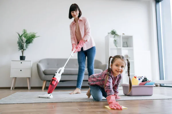Preteen kid in latex gloves cleaning floor from dirt while mother removing dust from carpet with canister vacuum. Tireless ladies of house reducing risk of allergies by taking care of home chores.